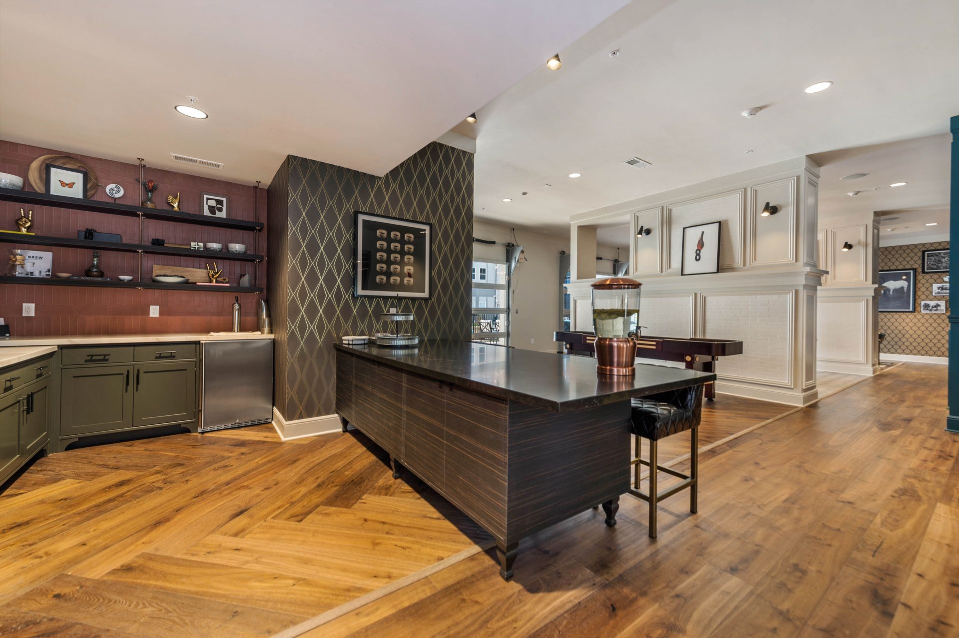 Contemporary kitchen area featuring a large dark countertop, green cabinetry, and decorative shelving with various items, set in a bright, open space with hardwood flooring.
