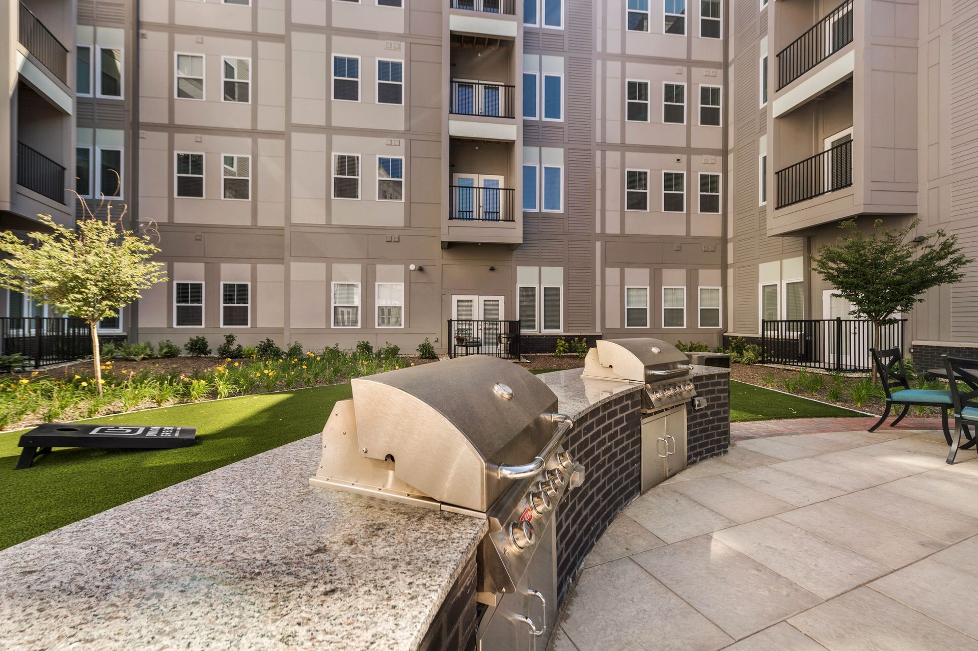 Outdoor grilling area with modern grills, surrounded by greenery and apartment buildings.