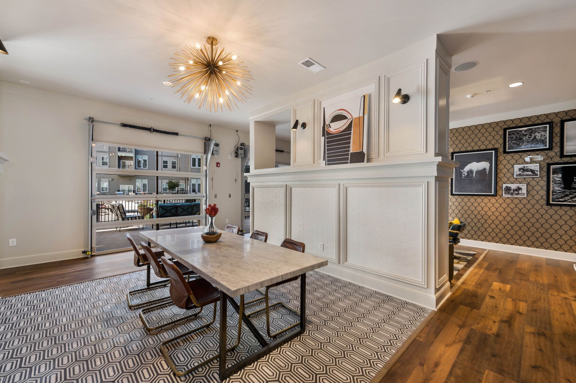A modern dining area featuring a marble table, stylish chairs, a decorative light fixture, and artwork, with large glass doors opening to an outdoor space.