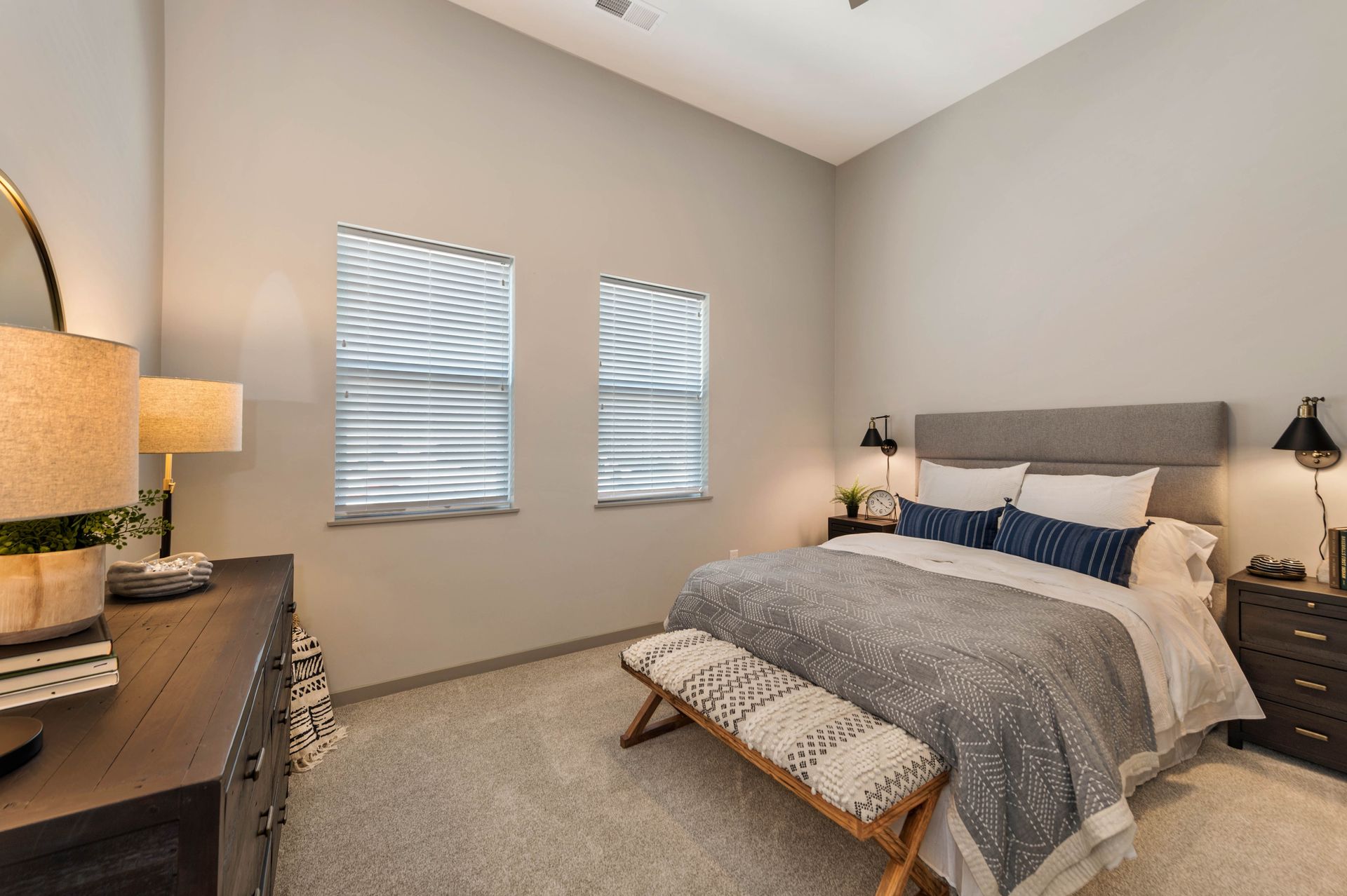 Cozy bedroom featuring a bed with gray and white bedding, two windows with blinds, a bedside table with a lamp, and a decorative bench at the foot of the bed.