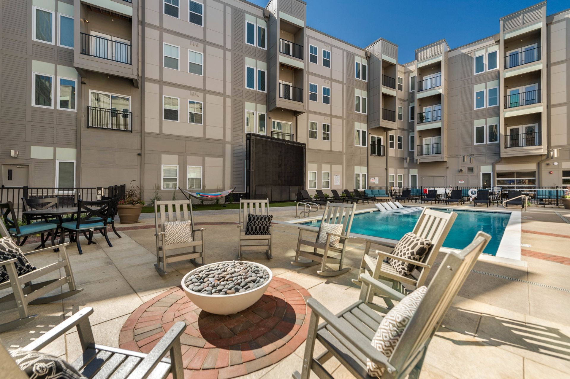A modern outdoor space featuring a pool surrounded by lounge chairs and a fire pit, with a multi-story building and hammocks in the background.