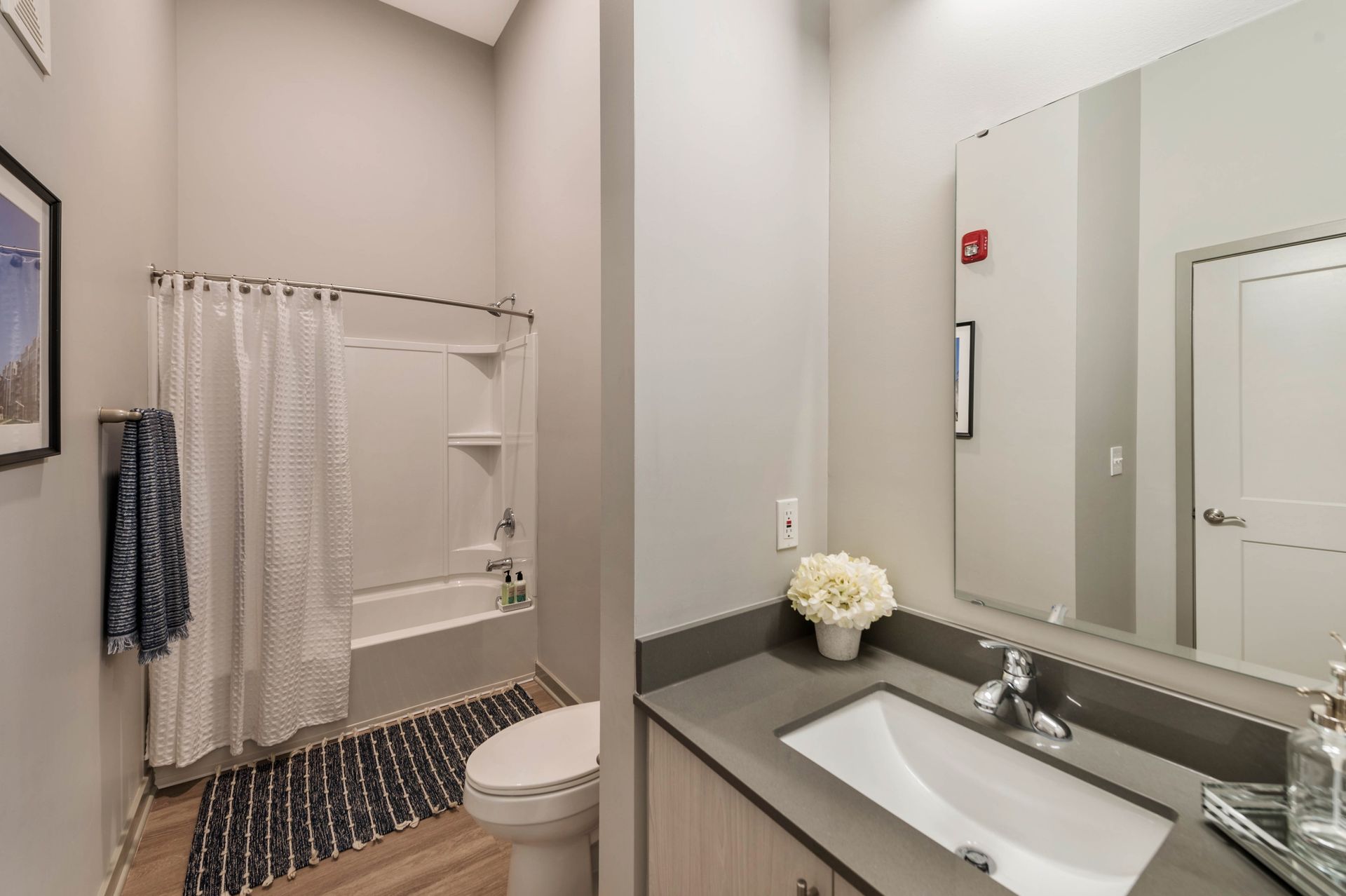 A modern bathroom featuring a shower with a white curtain, a toilet, and a sink with a gray countertop, along with decorative elements like a small vase of flowers and a dark patterned rug.