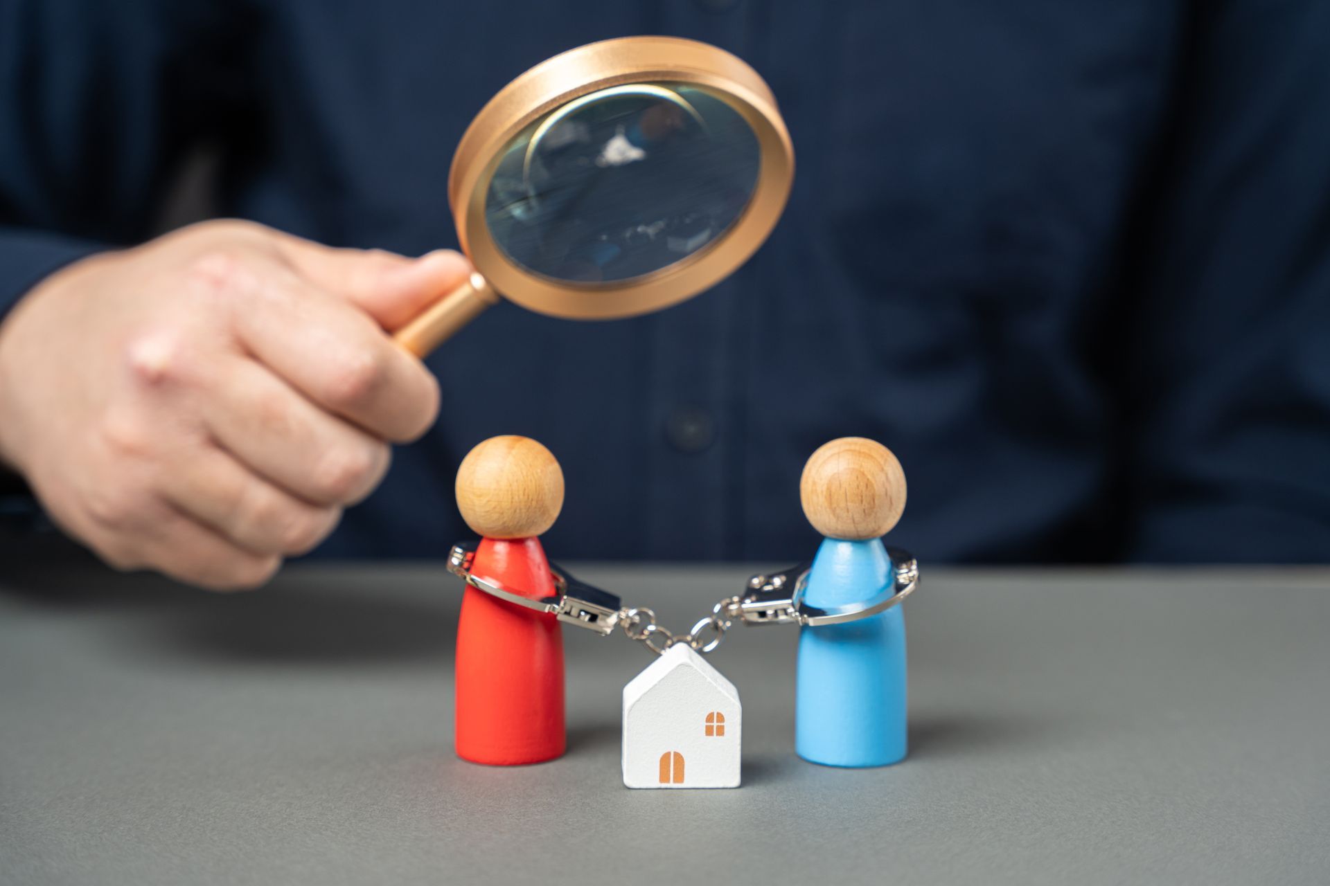 Person holding magnifying glass over two handcuffed figurines and a small house.