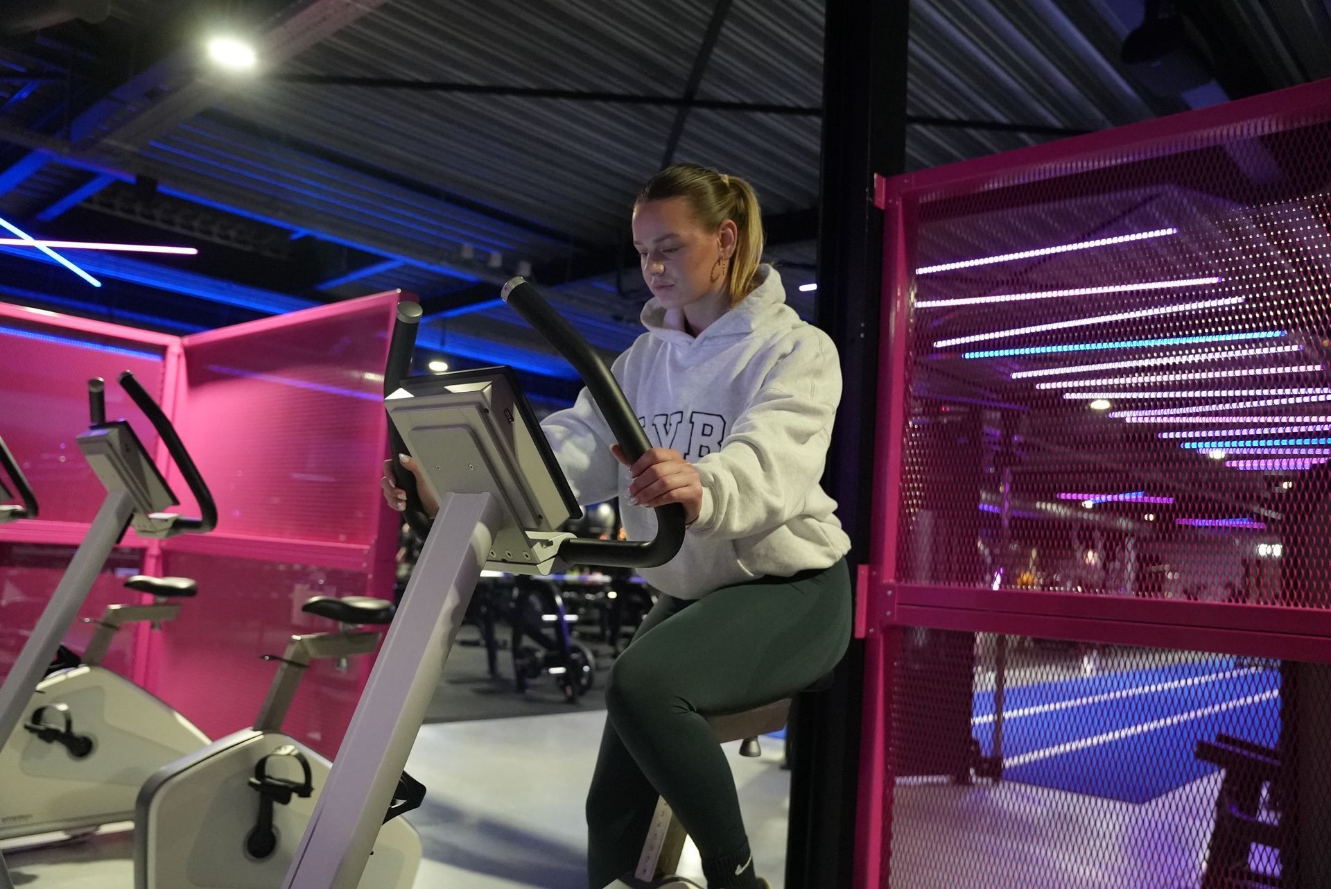 Woman in a gym on a stationary bike, wearing a hoodie, with pink accents.