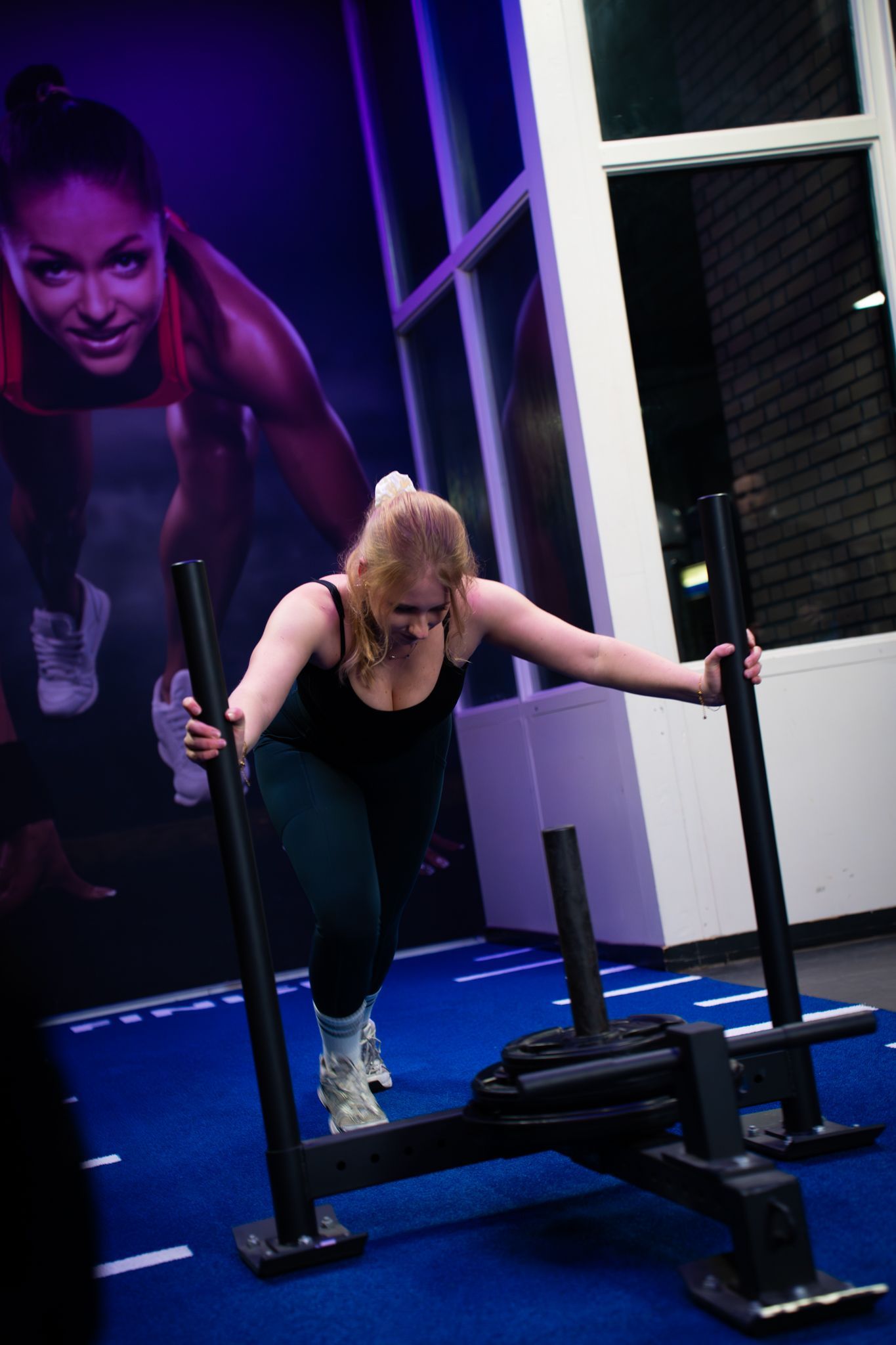 Een vrouw duwt een verzwaarde slee in een sportschool met paarse verlichting en fitnessapparatuur in de buurt.