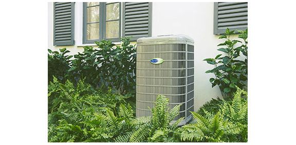 Outdoor air conditioner unit beside house windows, surrounded by green shrubs and plants