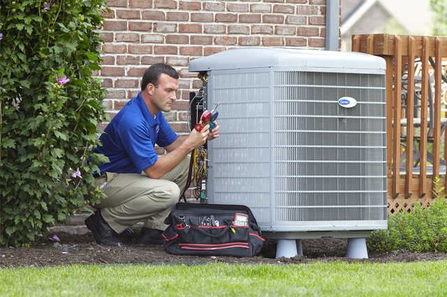 Technician kneeling beside an outdoor HVAC unit, using tools to inspect it in a yard.