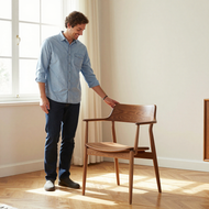 Man standing beside a wooden chair in a bright room, touching the chair’s backrest.