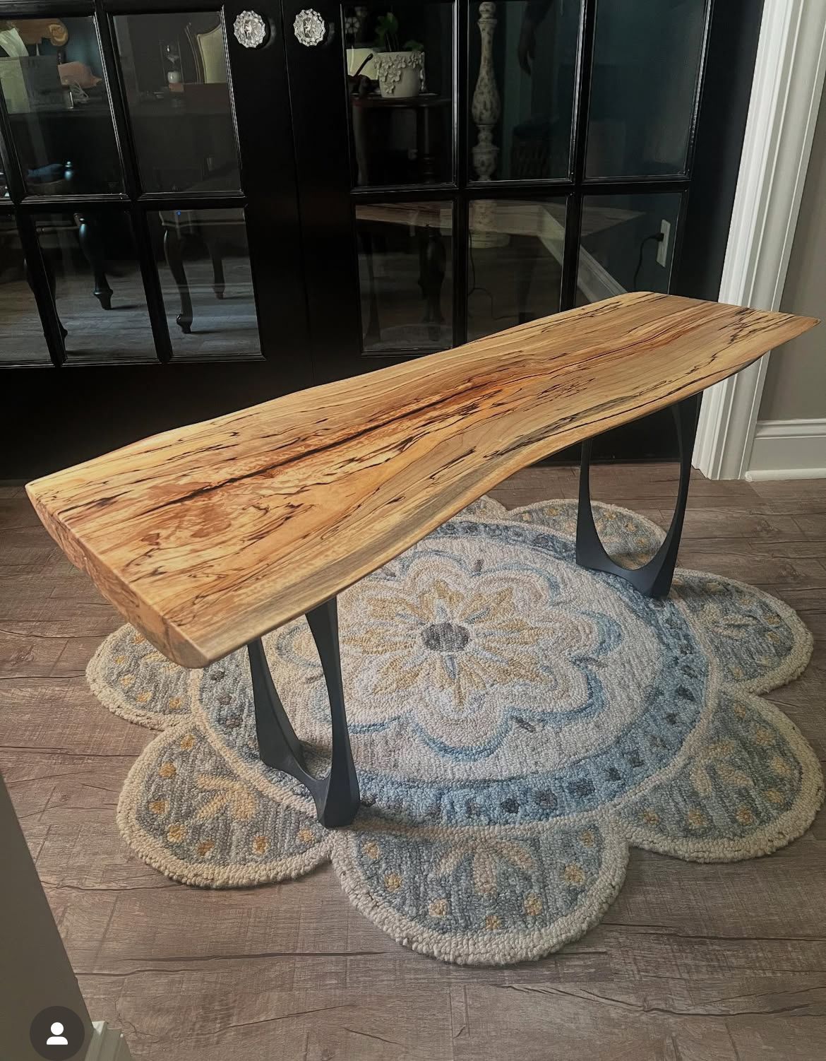 Live-edge wooden bench with black metal legs on a floral rug, set in front of black-framed glass doors.