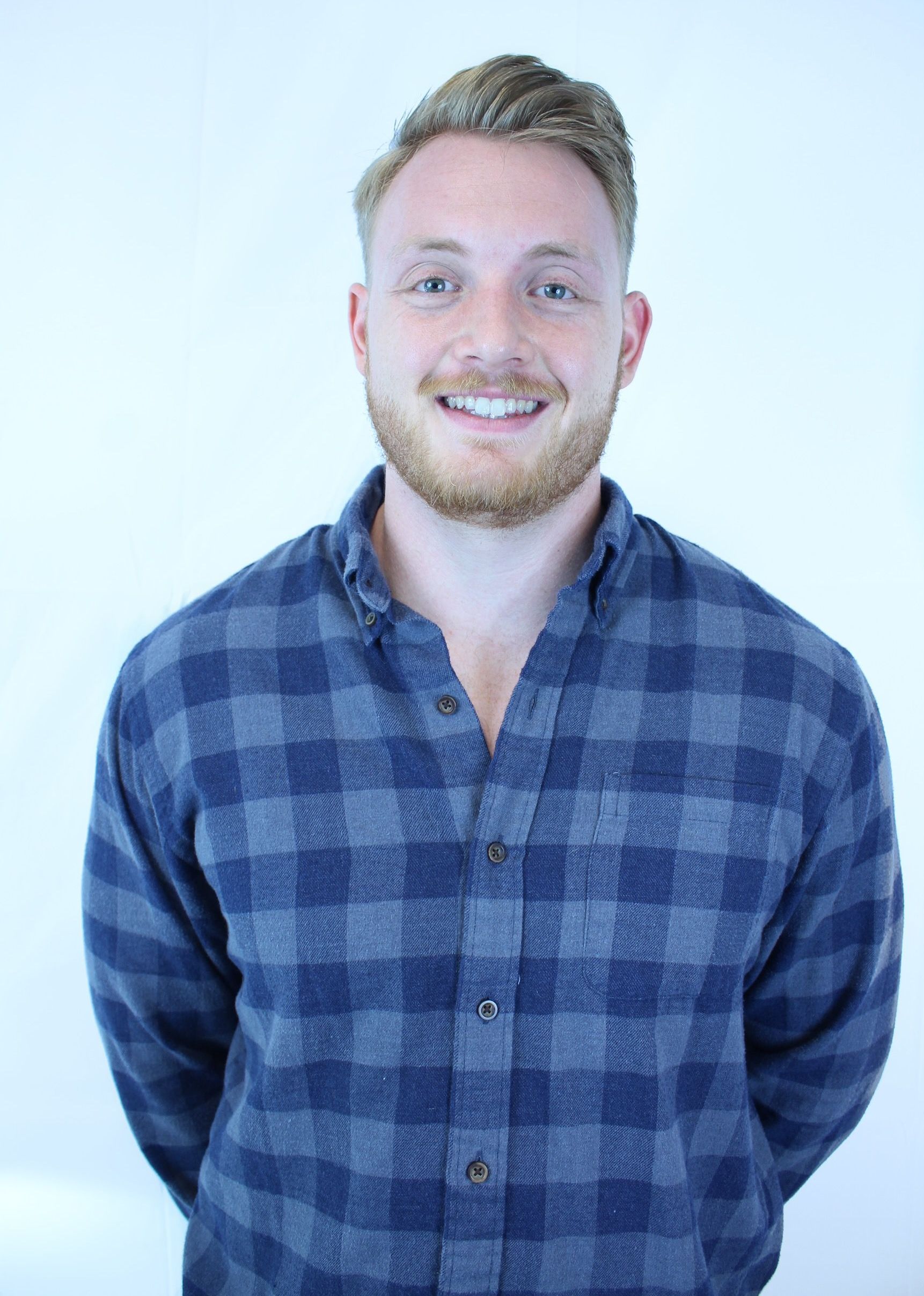 A man with a beard is wearing a blue plaid shirt and smiling.