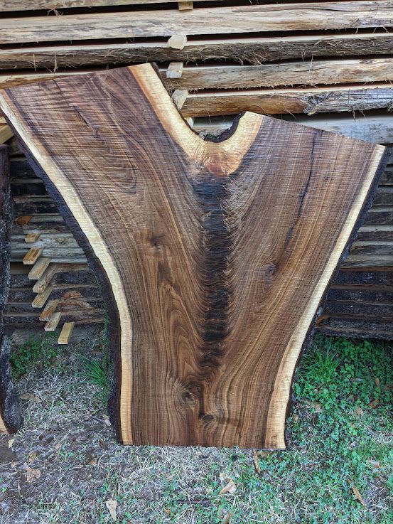 Live edge walnut slab with a Y-shaped grain pattern, surrounded by lighter sapwood. Stacked lumber in the background.