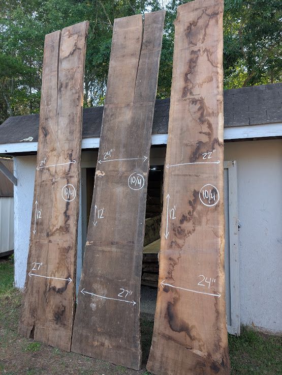 Three long, rough-cut wooden slabs leaning against a white shed in an outdoor setting. Each slab has chalk markings.