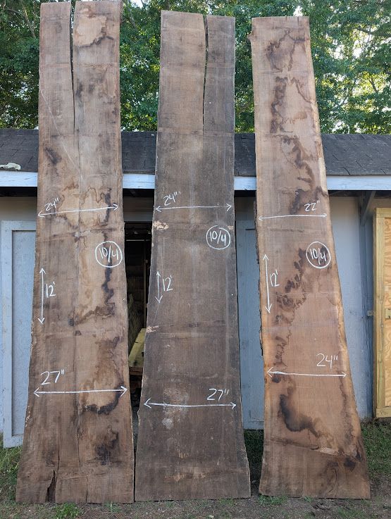 Three long, unfinished wooden planks stand against a building. White measurements and numbers are marked on the wood.