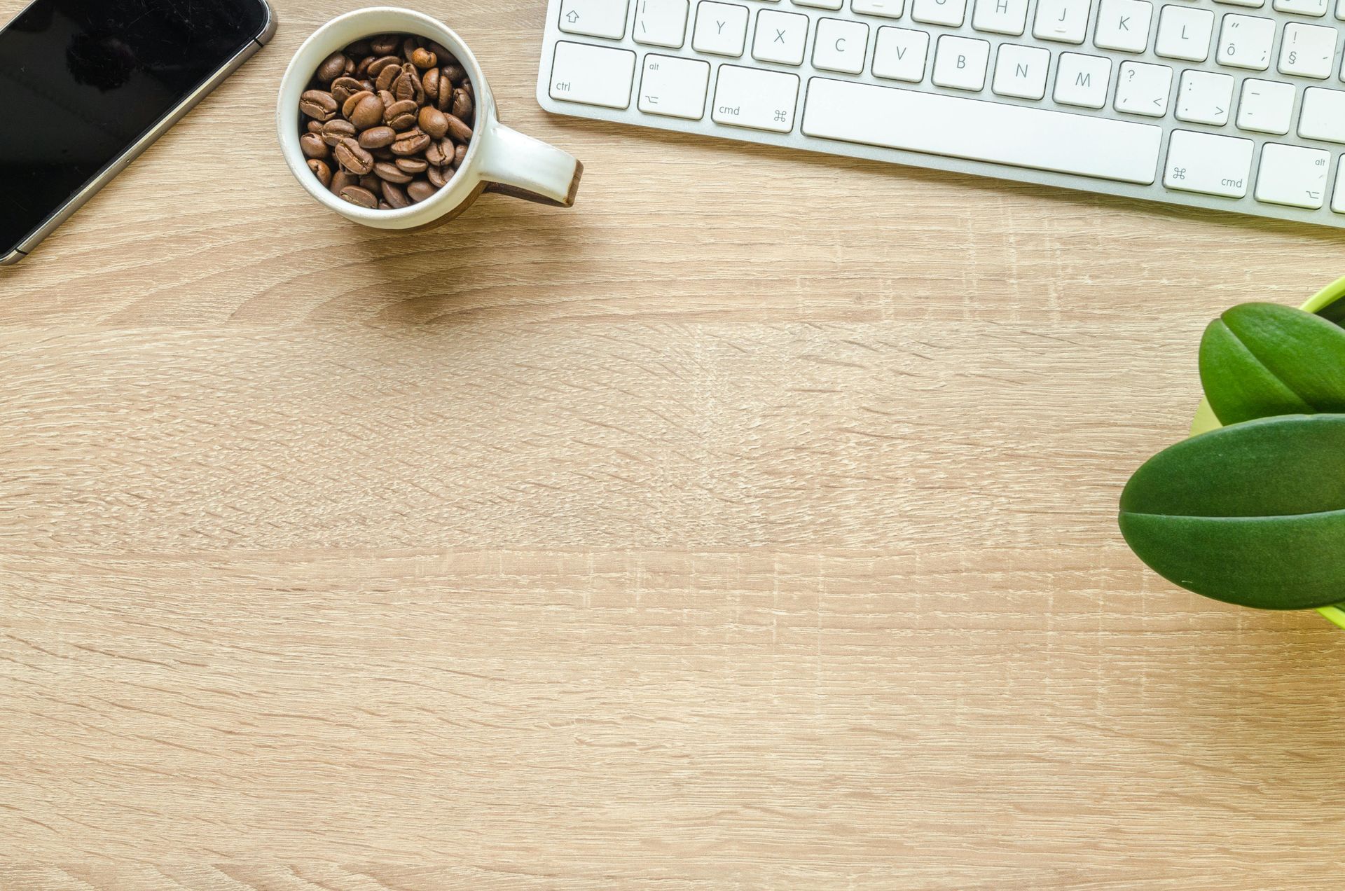 Overhead shot of a wooden desk with a keyboard, coffee beans in a mug, a phone, and a potted plant.