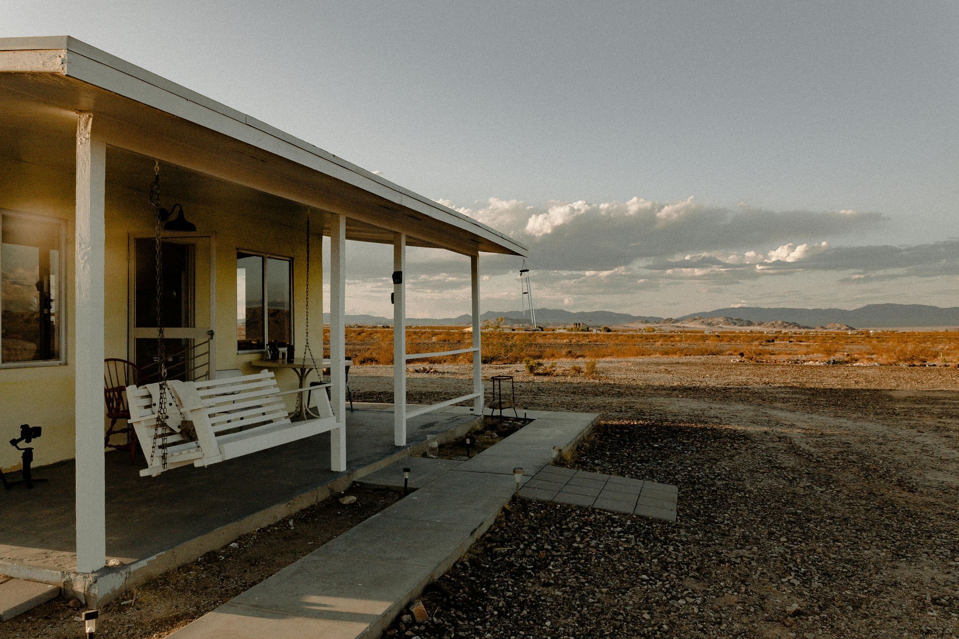 Yellow house with a porch swing overlooks a vast, dry landscape under a cloudy sky.