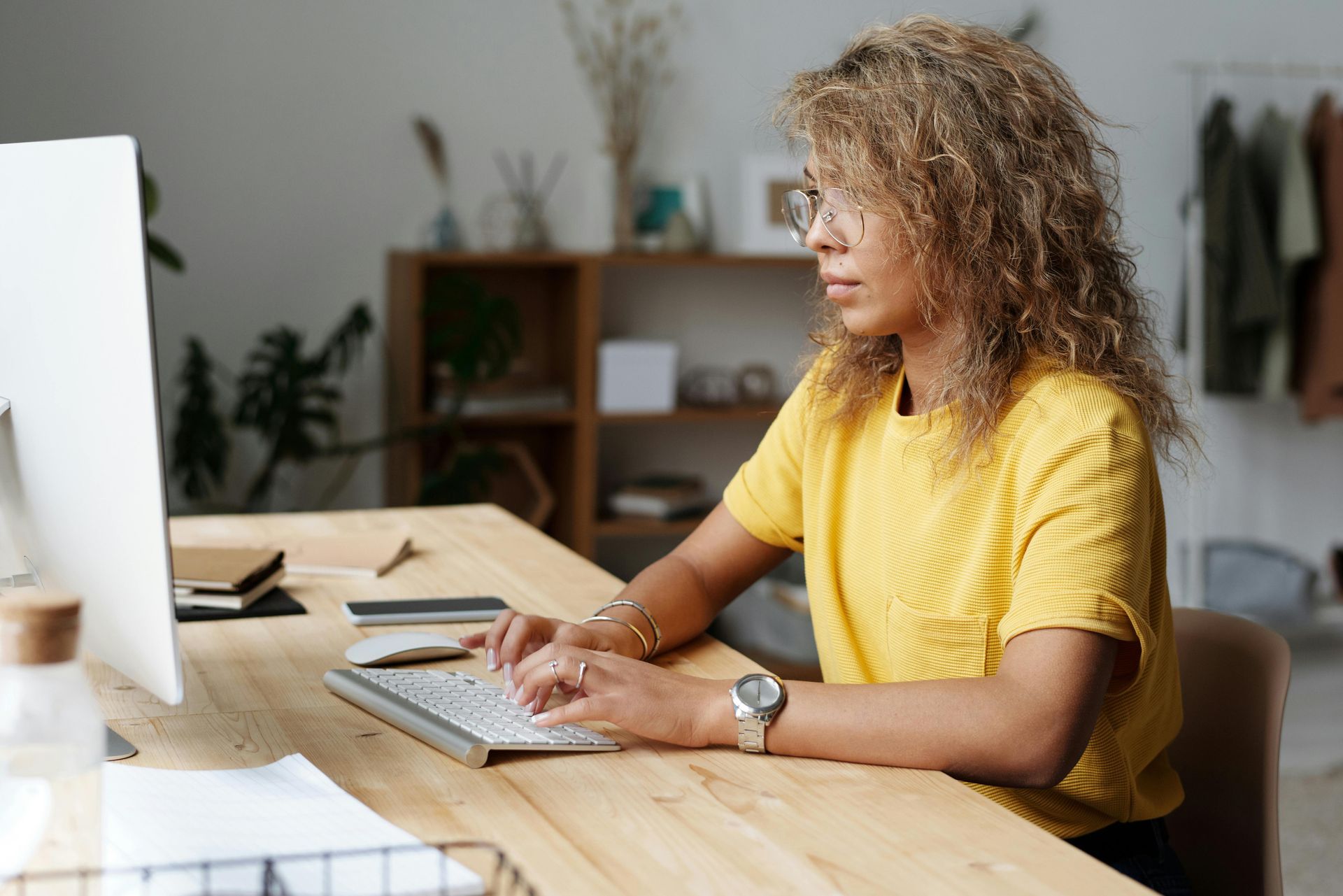 Woman with curly hair wearing glasses and a yellow shirt working on a computer at a wooden desk in a well-lit room.