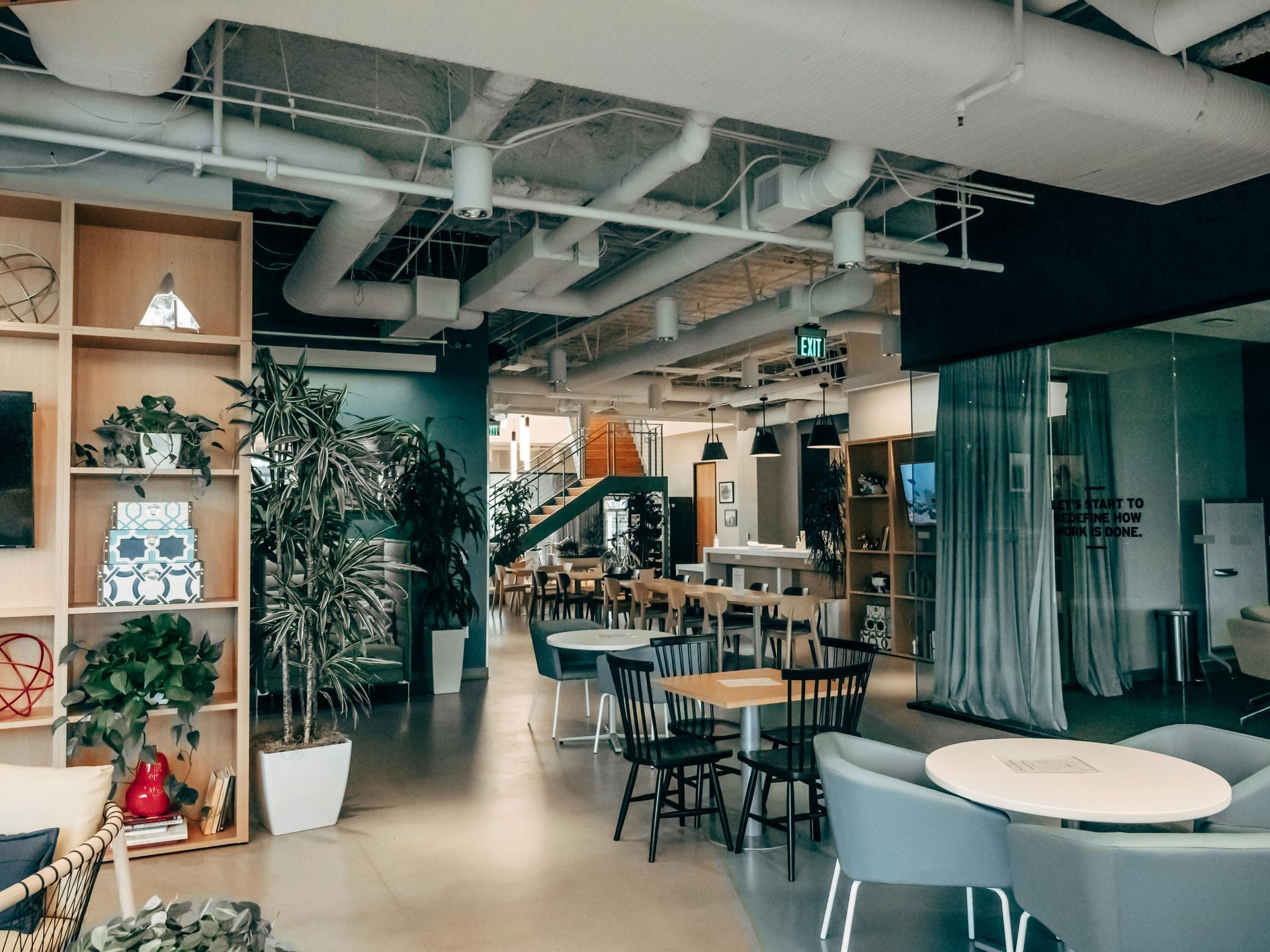 Modern open-plan office with tables, chairs, plants, and exposed pipes on the ceiling.