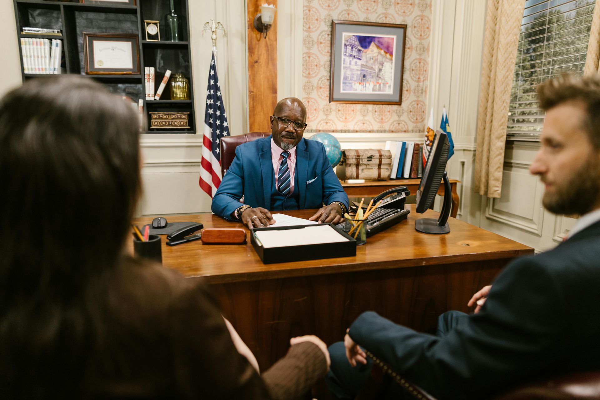 A Black man in a blue suit sits at a desk, facing two people. An American flag and artwork are in the background.