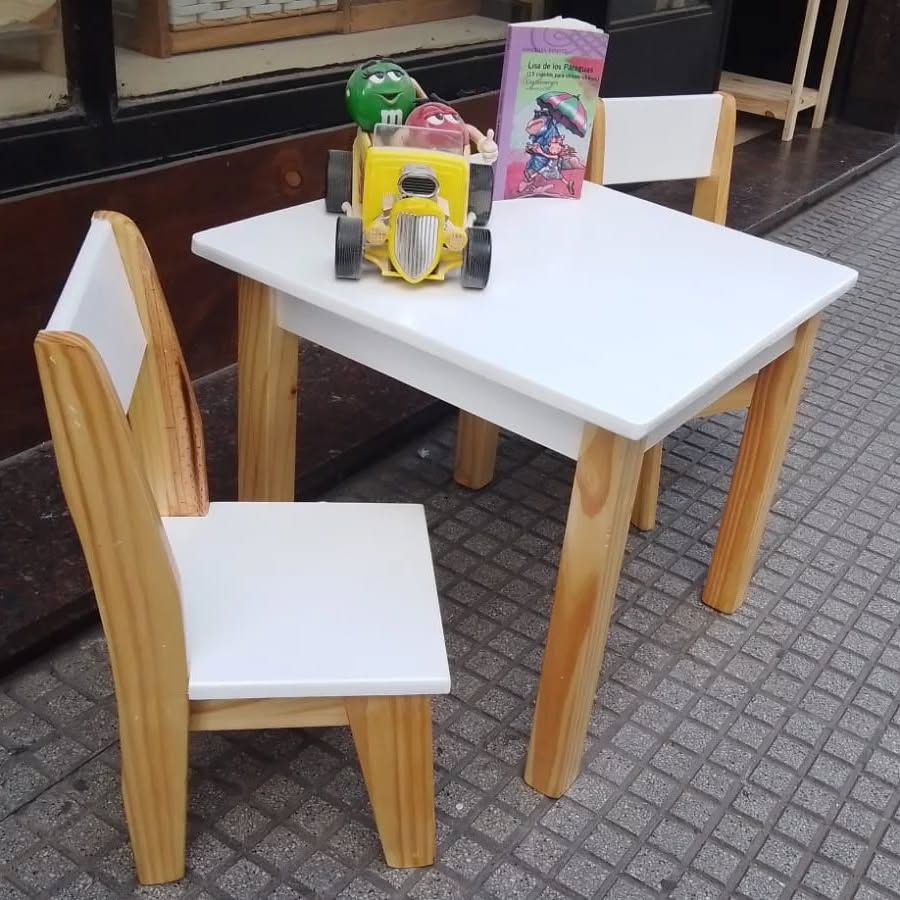 Mesa infantil blanca y dos sillas con patas de madera. Hay juguetes sobre la mesa, en la acera.