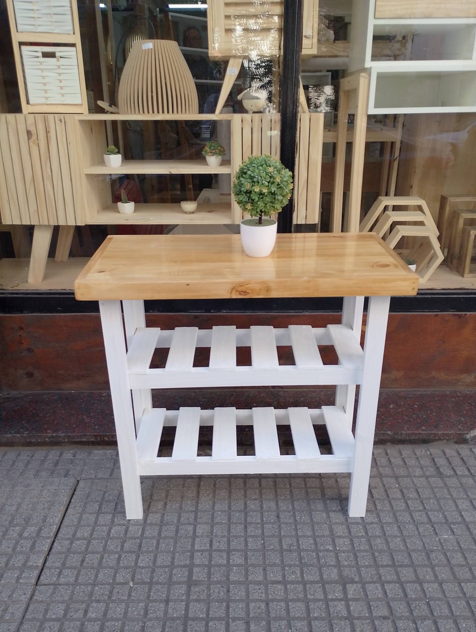Mesa de madera con base blanca y tablero marrón claro, situada sobre una acera de baldosas, con elementos decorativos en el fondo.