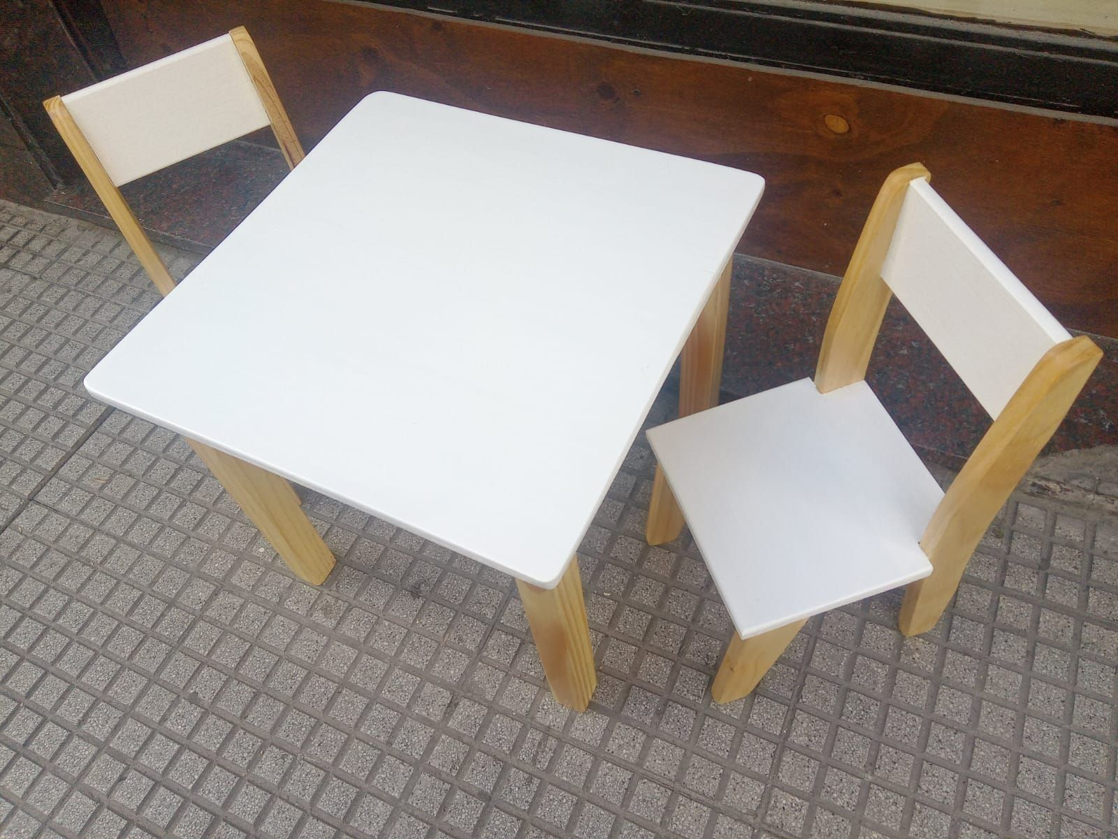 Mesa infantil cuadrada blanca y dos sillas en una acera. Los asientos de la mesa y las sillas son blancos; las patas son de madera clara.