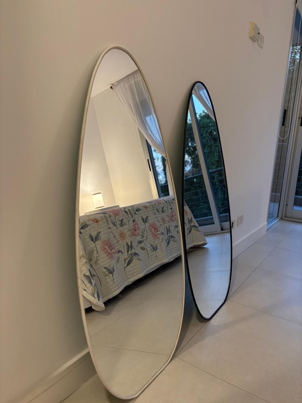 Dos espejos ovalados apoyados sobre una pared blanca, uno sin marco y otro con marco negro, que reflejan un dormitorio con una cama.