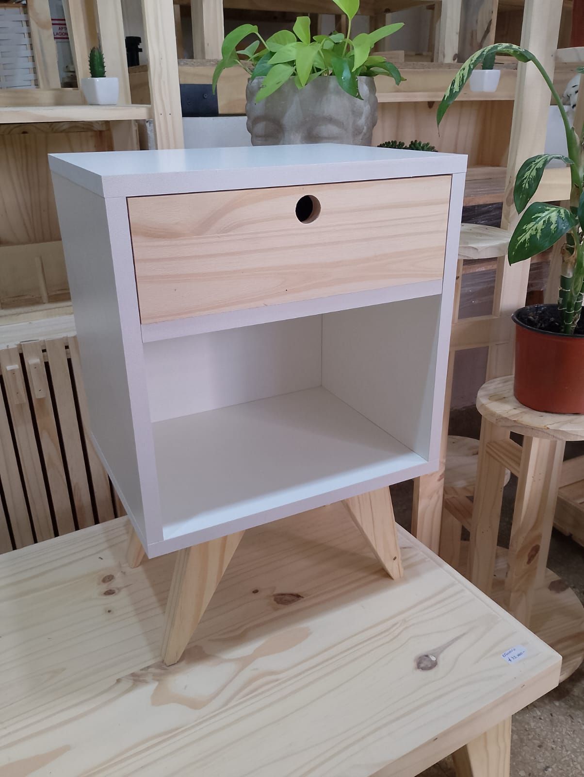 Mesita de noche de madera y color blanco con cajón y estante, patas en ángulo, sobre fondo rosa.