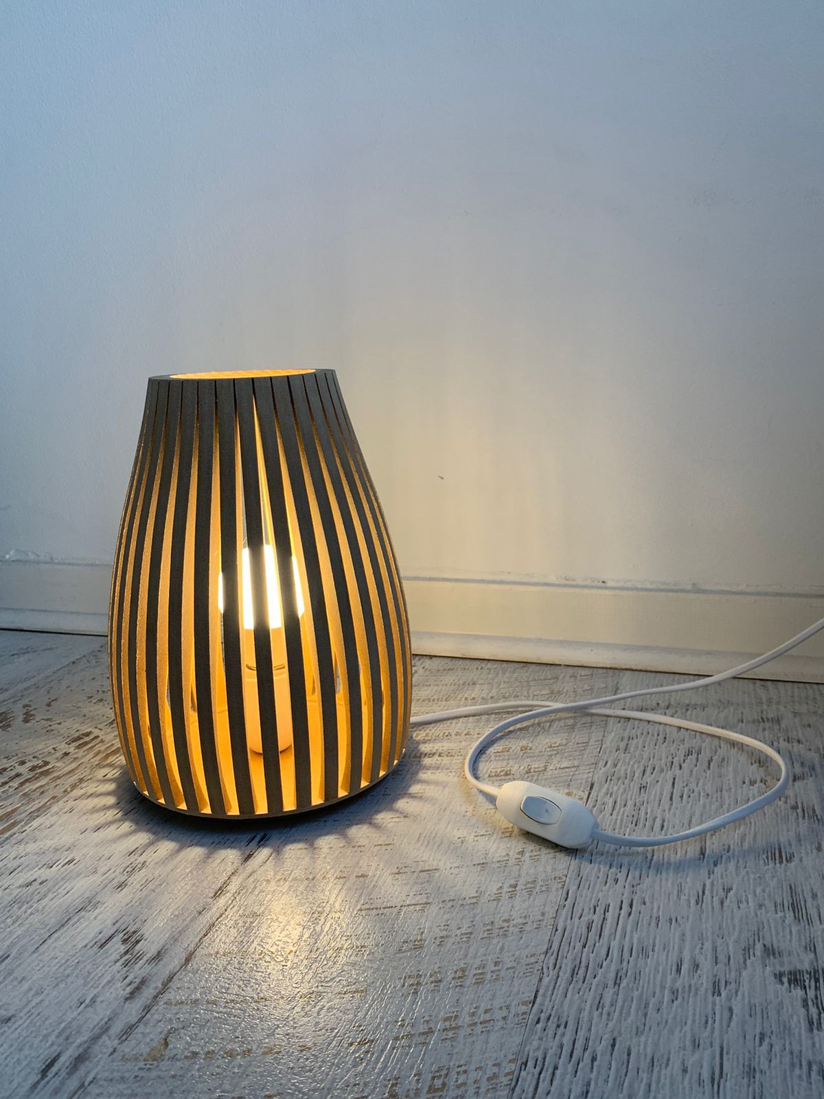 Lámpara de madera con ranuras verticales que proyectan luz y sombra sobre el suelo y la pared.