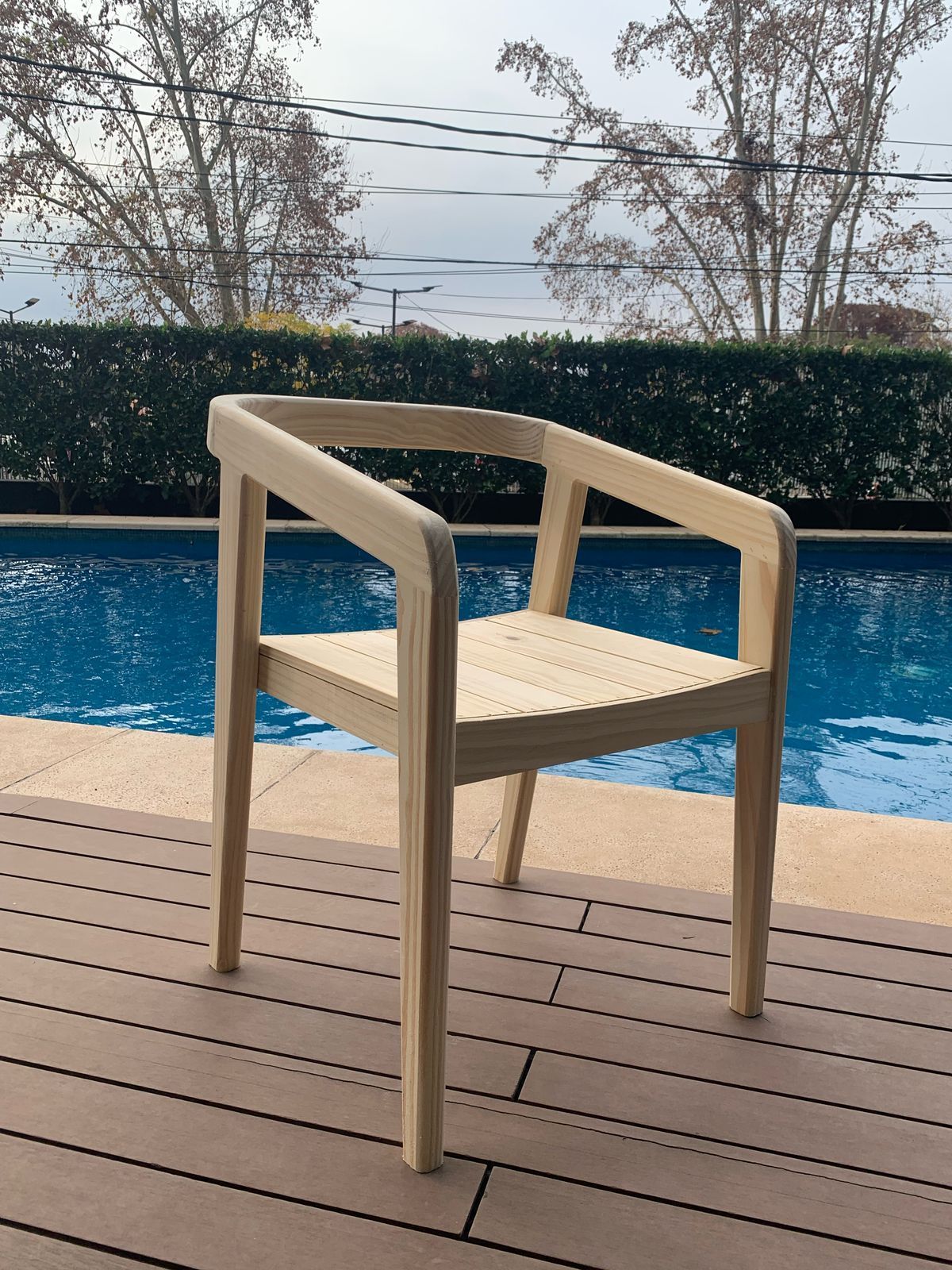 Silla de madera con brazos, madera de color claro, sobre un patio de ladrillo, con plantas verdes al fondo.