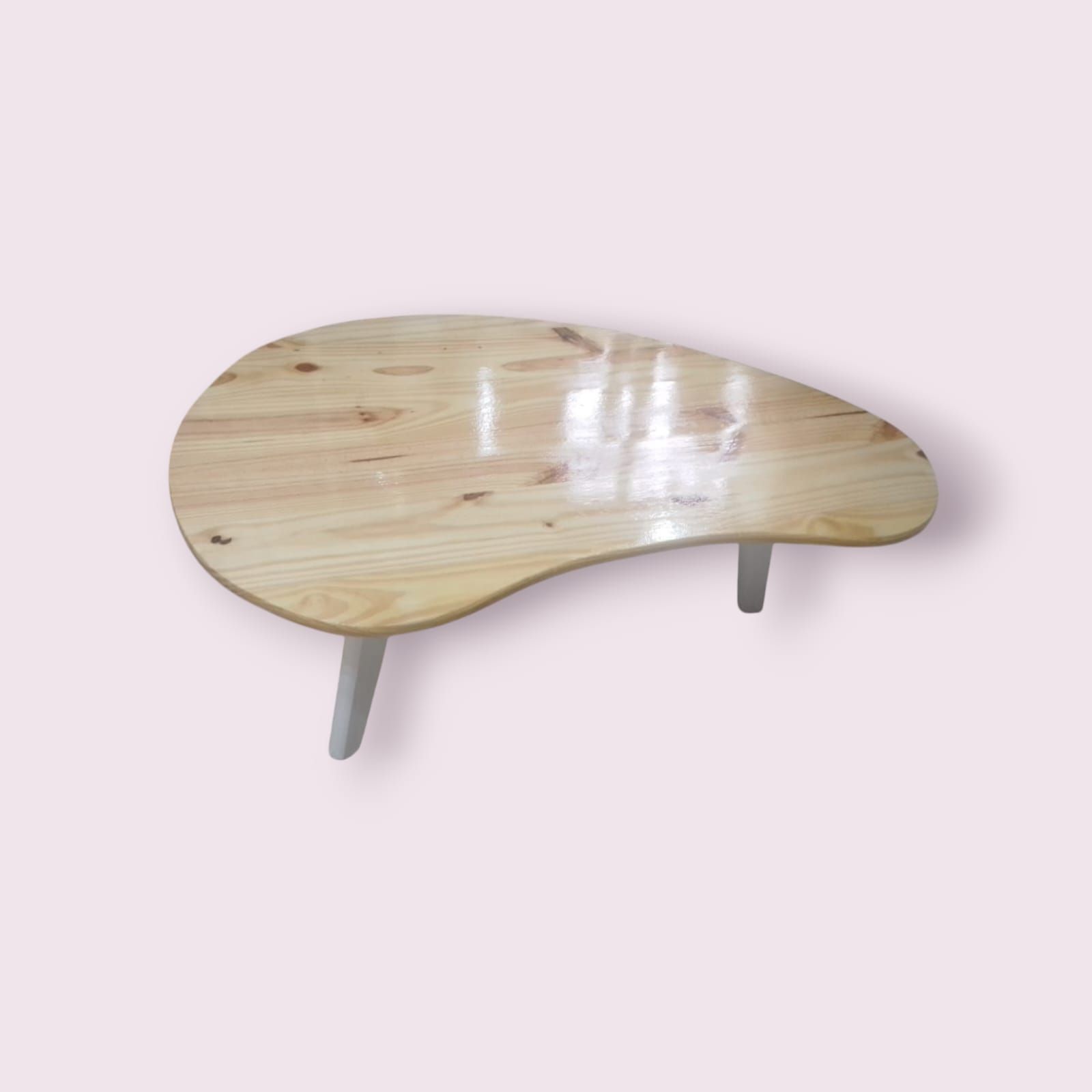 Mesa de centro en forma de lágrima, de color madera clara, con patas blancas sobre un fondo rosa.