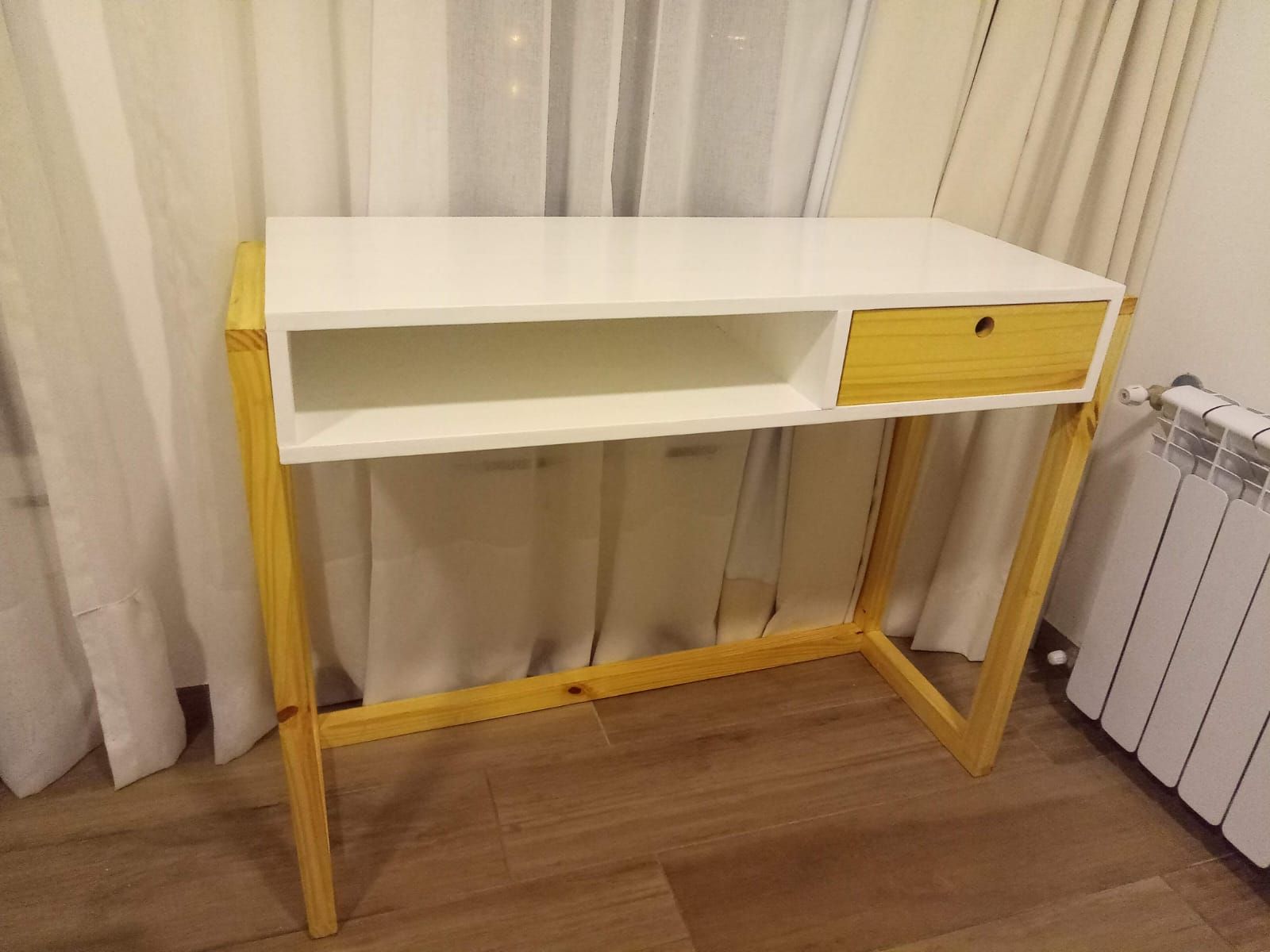 Escritorio blanco y de madera con cajón y estante abierto, sobre un fondo de cortina blanca.