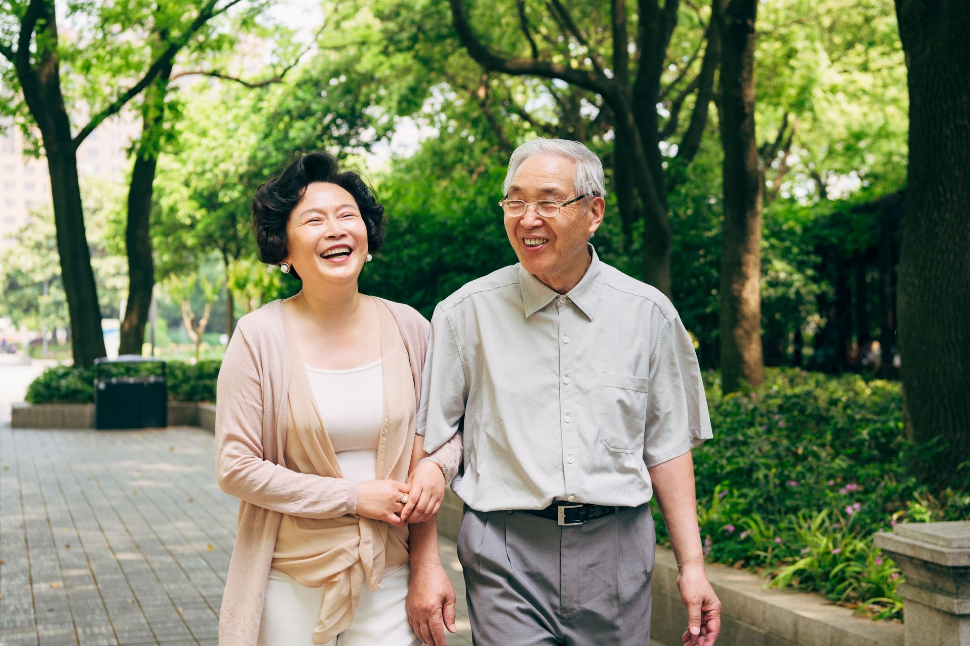 An elderly couple is walking down a sidewalk in a park.