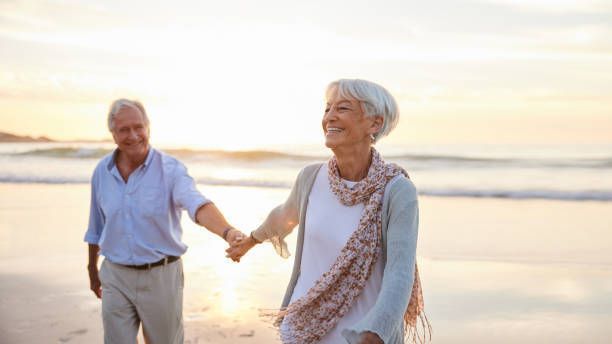 An elderly couple is walking on the beach holding hands.