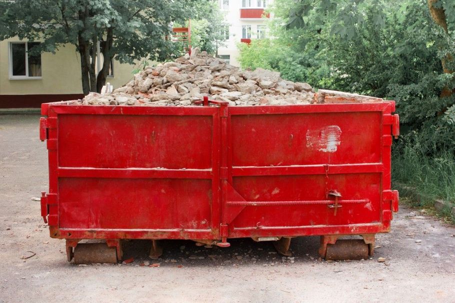 Red dumpster filled with rubble on a dirt road, with trees and a building in the background