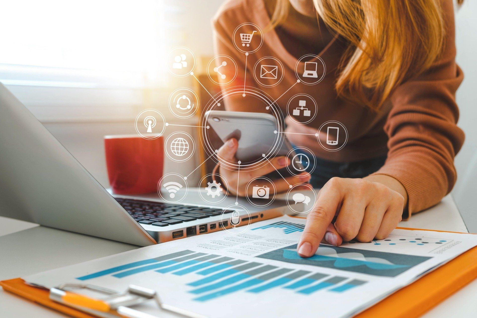 Person mit Kaffee und Smartphone blättert in einem Katalog, Laptops und Unterlagen auf dem Tisch
