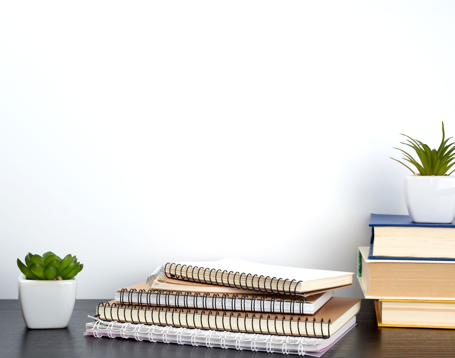 Desk with stacked notebooks, books, and small potted succulents against a white wall.