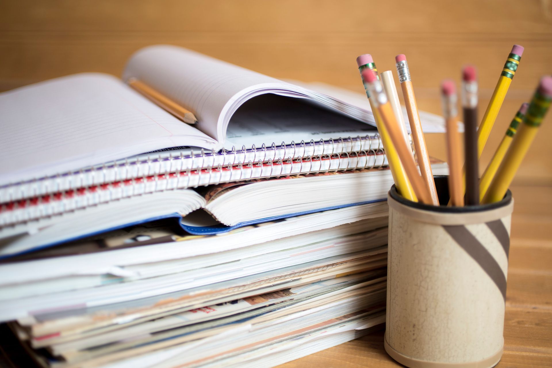 Stack of notebooks and papers with pencils in a container on a wooden surface.