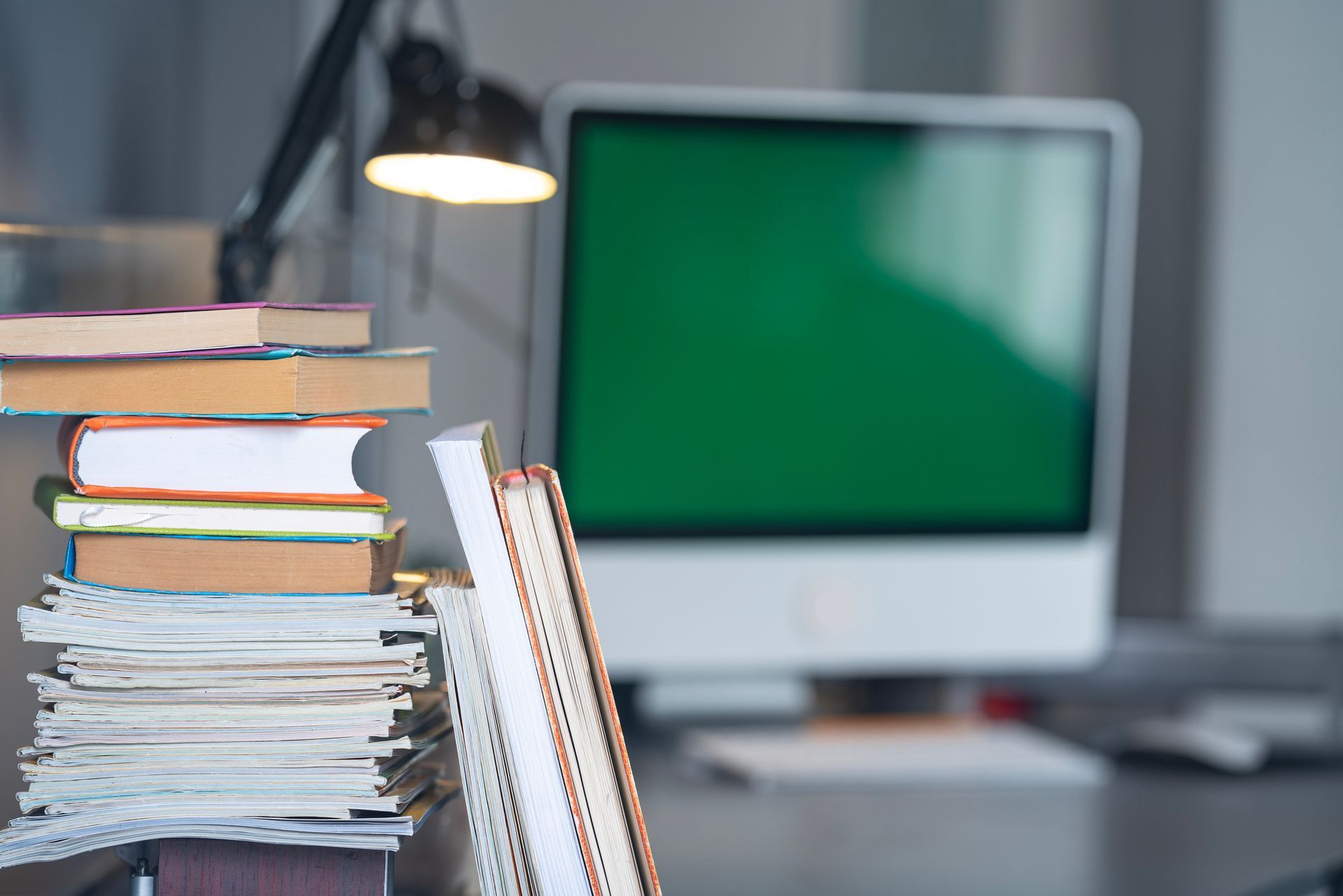 Stack of books and papers with a lamp and computer, which has a green screen, on a desk.