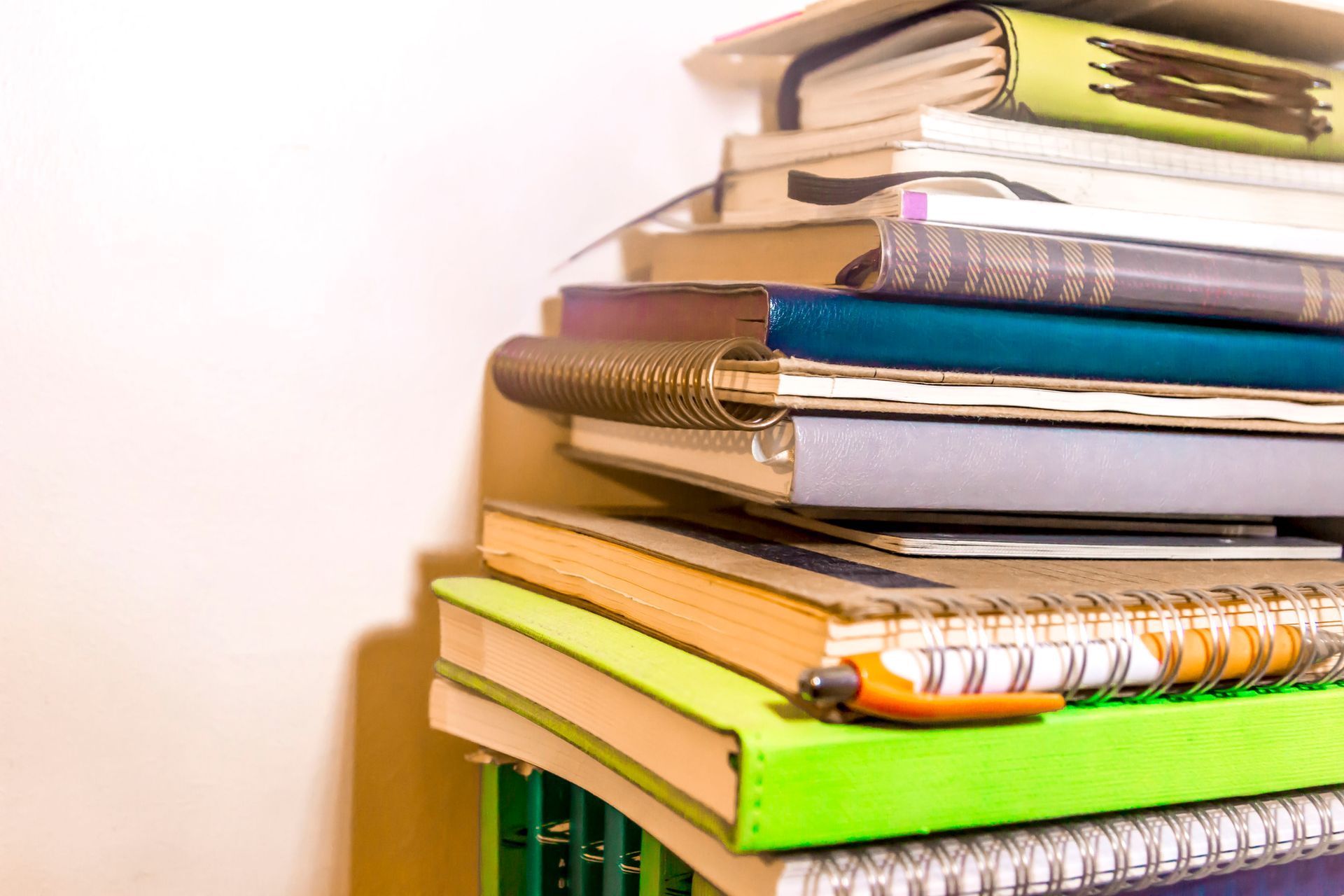 Pile of notebooks and books, various colors, with a pen resting on top.