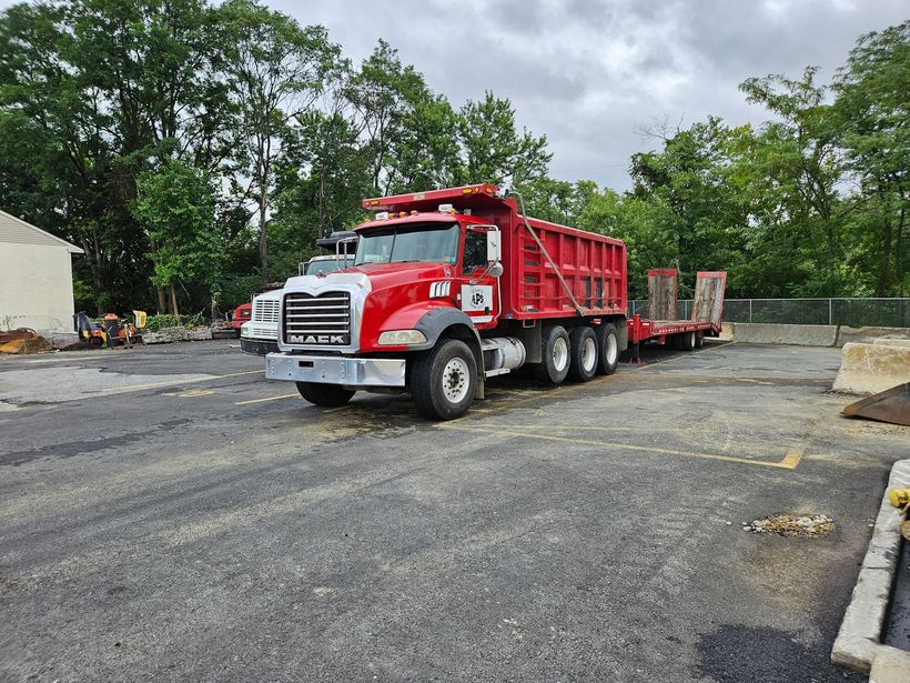 Red Mack dump truck with a trailer parked on gravel in front of trees.