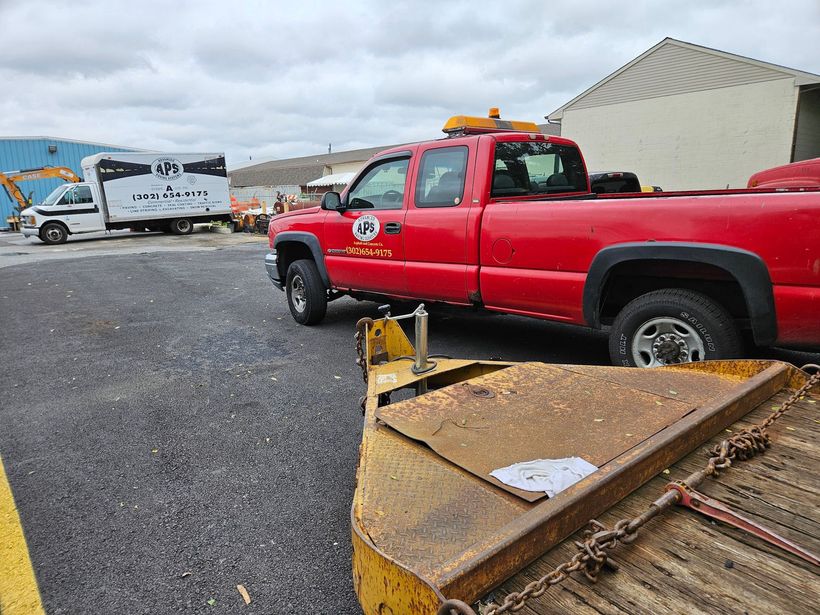 Red pickup truck with a snowplow and a white work truck on a grey asphalt lot in front of a building on an overcast day.