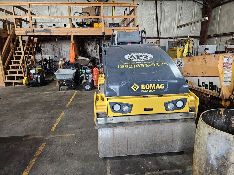 A yellow BOMAG road roller in a garage, next to a barrel. Other equipment is visible in the background.
