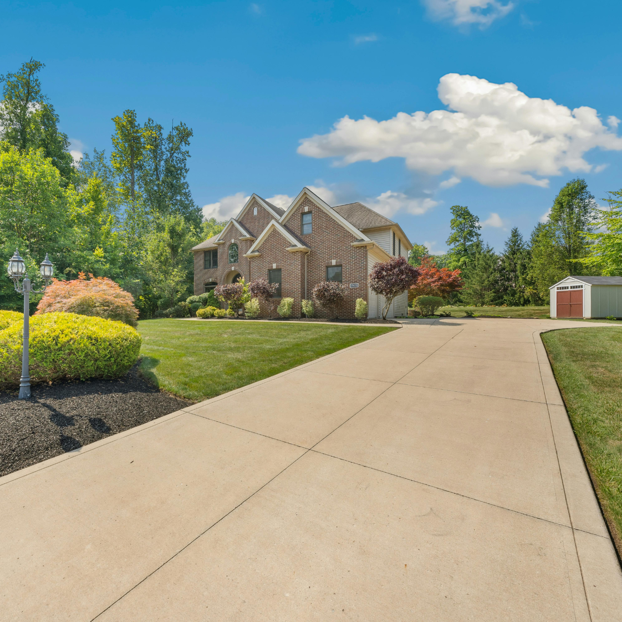 Brick house with long driveway, green lawn, and trees under a blue sky with clouds.