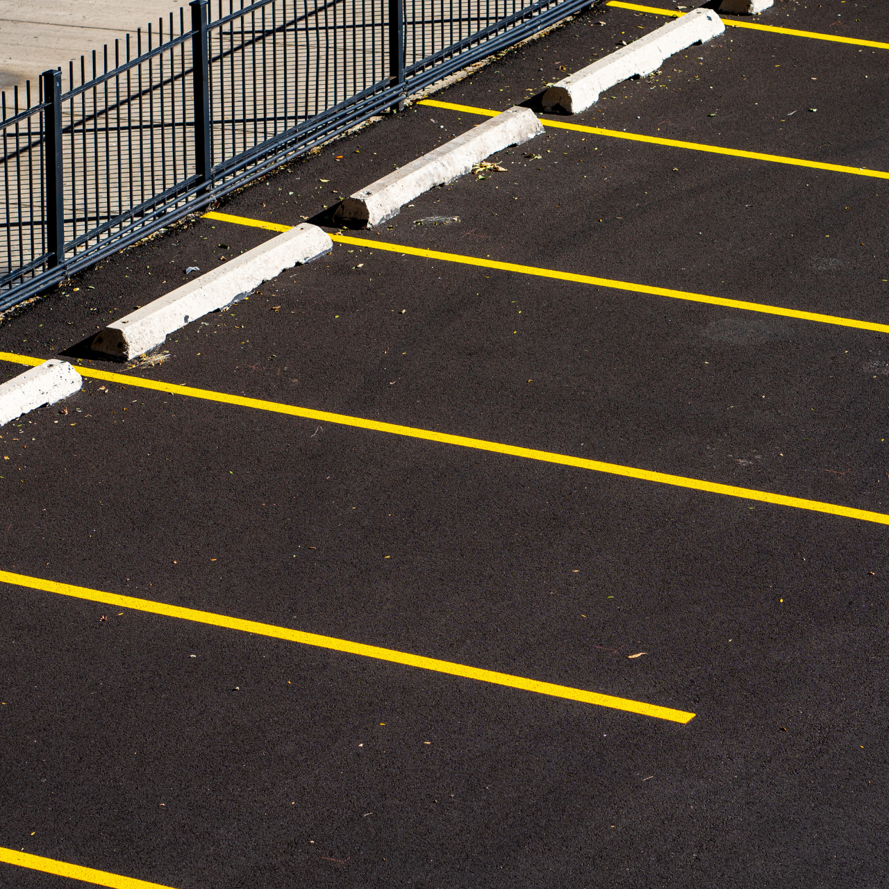 Empty asphalt parking lot with yellow lines and concrete wheel stops next to a metal fence.