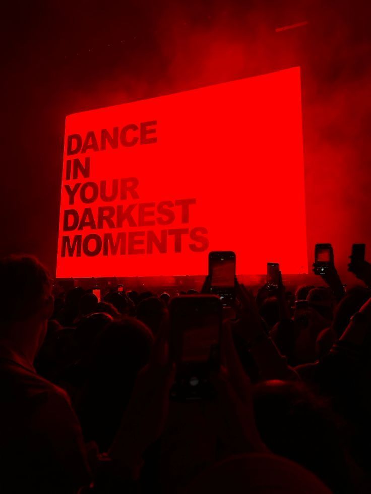 A crowd of people are standing in front of a red sign that says `` dance in your darkest moments ''.