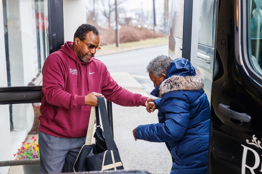 A person in a maroon hoodie helps an older adult board a van, holding a black bag for them.