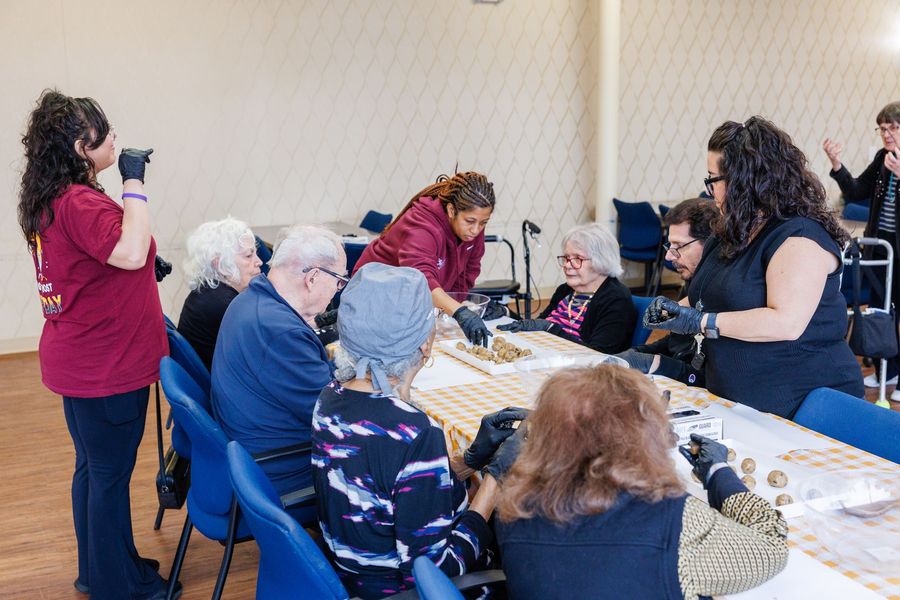 A food cart holds rows of bowls with pudding, while a person in a maroon shirt prepares to serve food in a dining room.