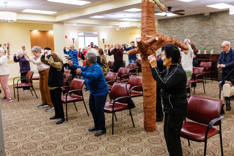A group of people standing in a community room, participating in a group fitness class with arm movements.
