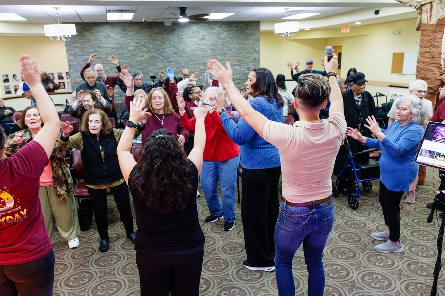 A group of people standing in a community room with their arms raised, following an instructor leading an exercise class.