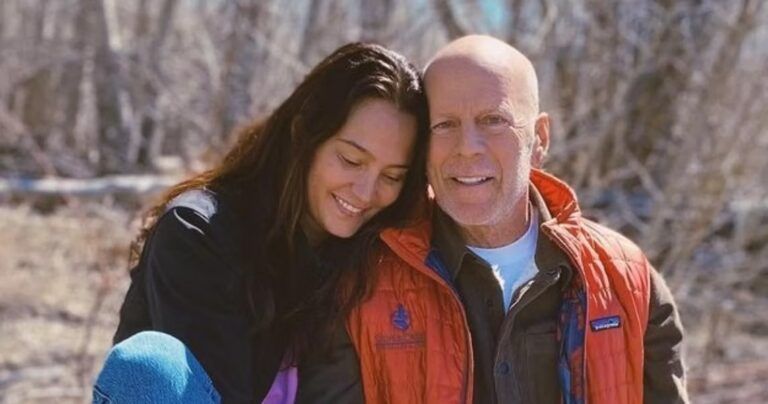 A woman leans her head against a man's head outdoors; both are smiling in a wooded, sunlit setting.